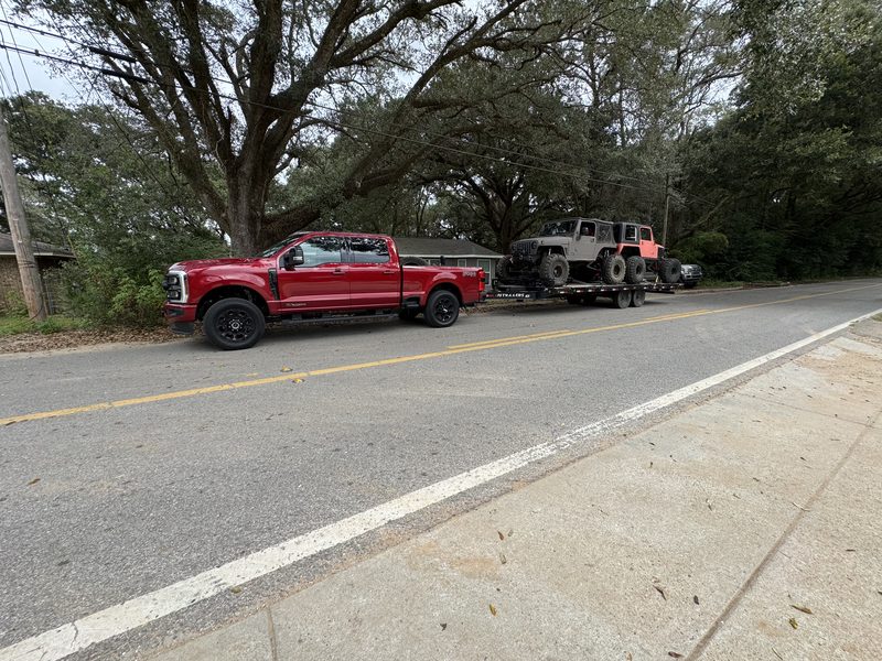 A deckover trailer carrying a Jeep body behind a pickup on a tree-lined Baldwin County road