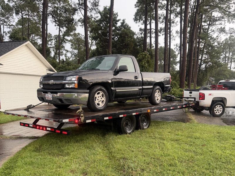 A full-size Chevy pickup strapped down on the deckover tilt trailer in a driveway